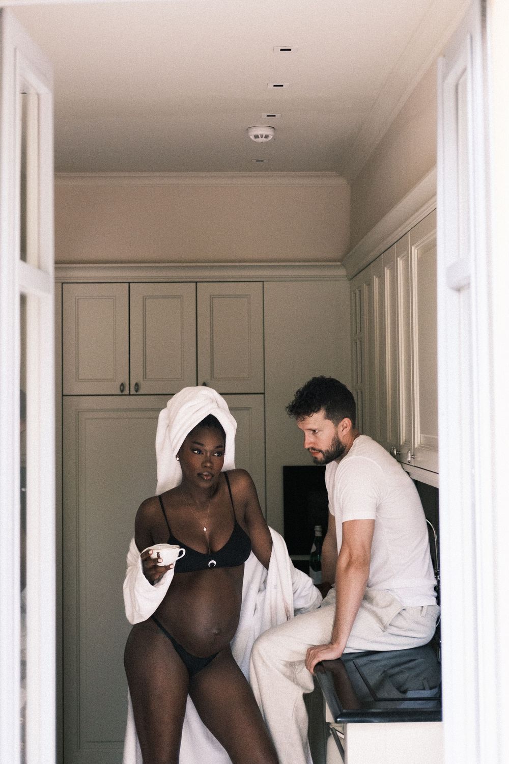 Pregnant Black woman in lingerie and white robe holds coffee cup while man sits on counter in modern kitchen.