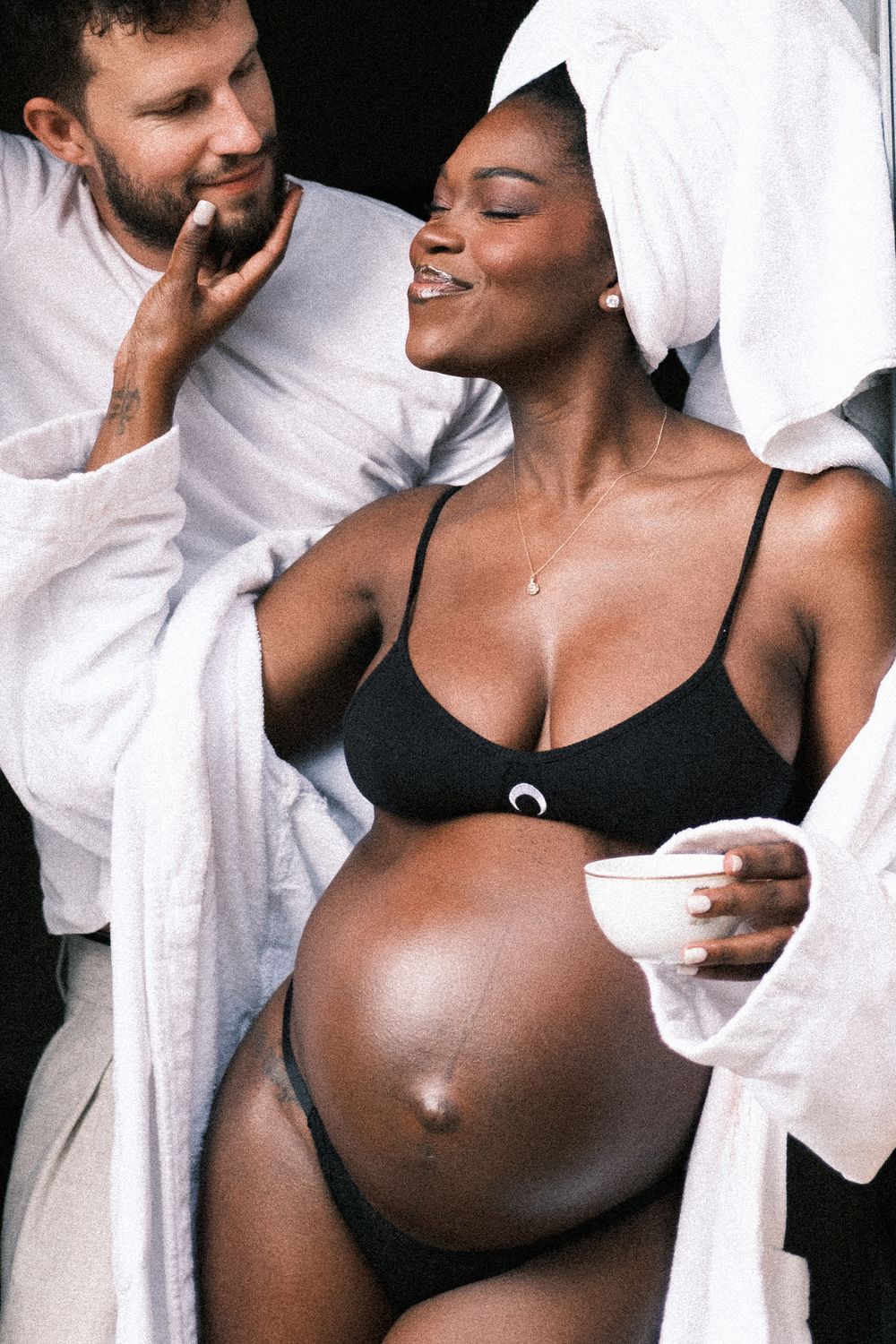 Pregnant woman in black lingerie with towel turban smiles holding cup beside man in white robe.