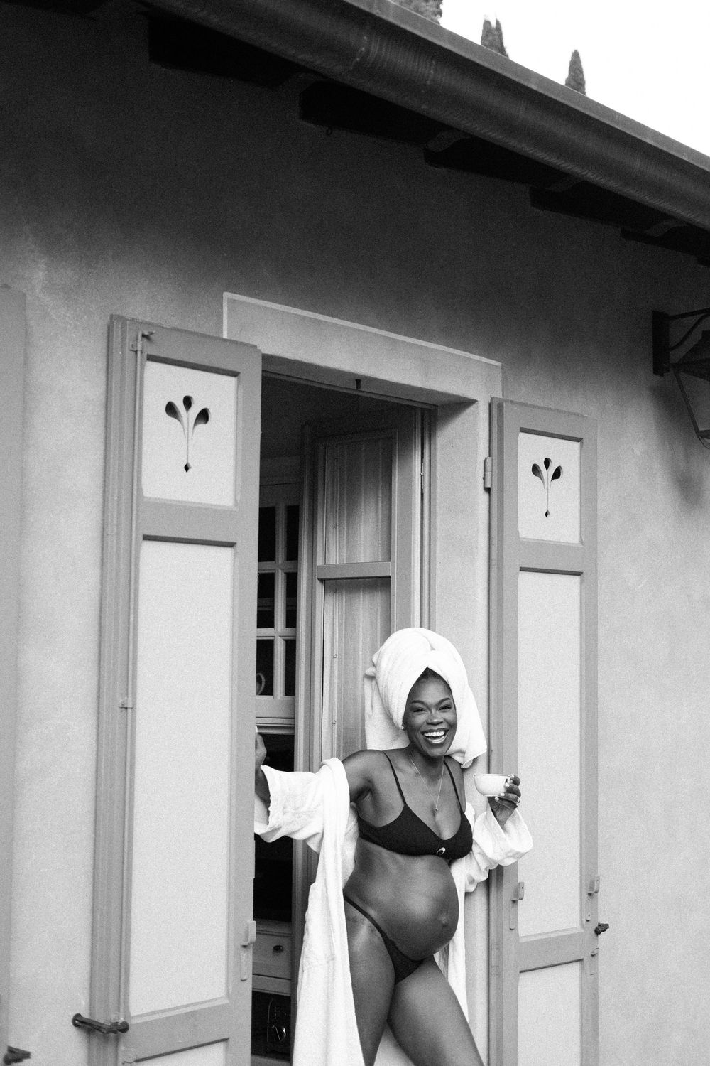 Woman in bikini and towel turban laughs joyfully in doorway of rustic building, black and white photo.