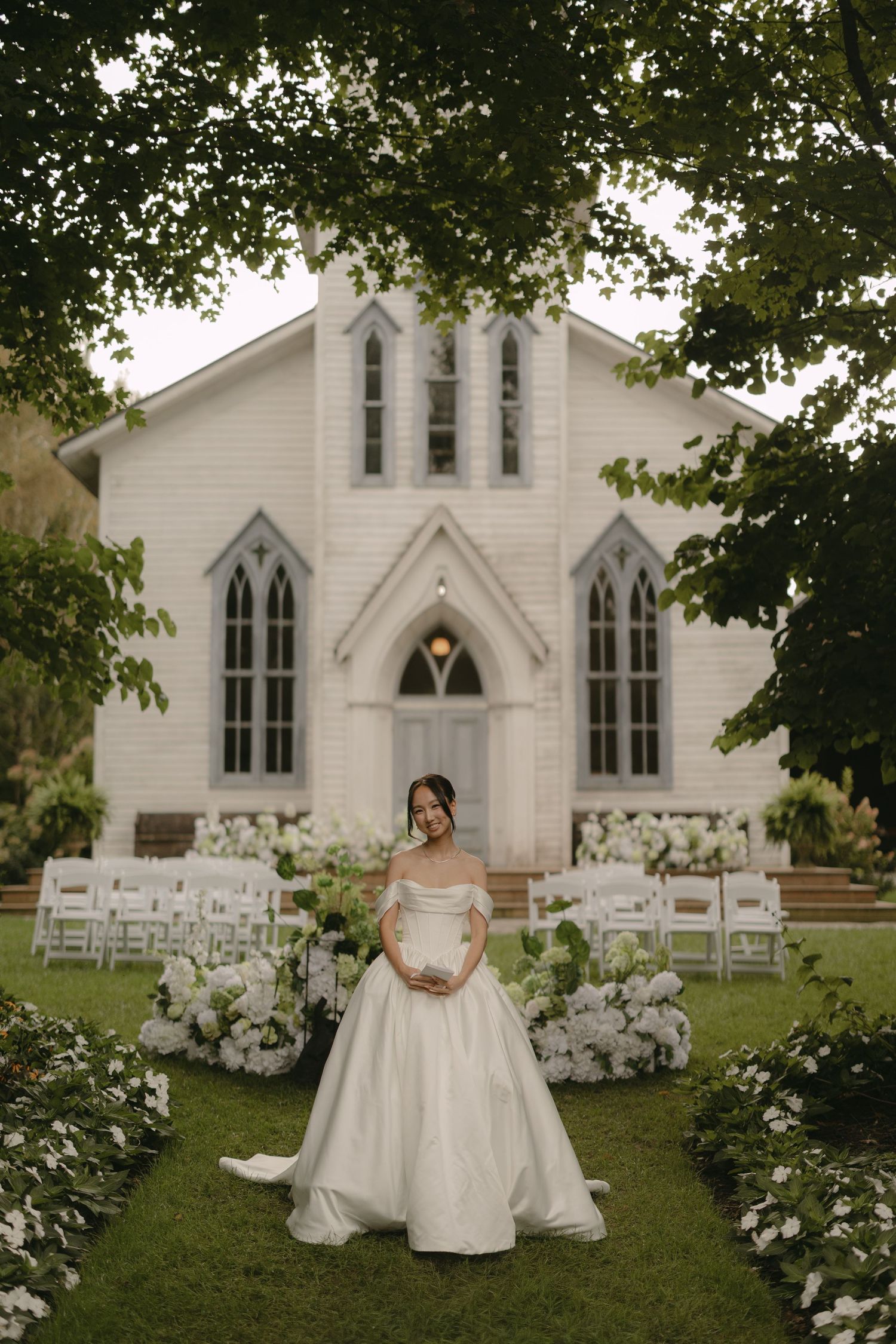 Bride in off-shoulder ball gown stands before white chapel decorated with white floral arrangements.