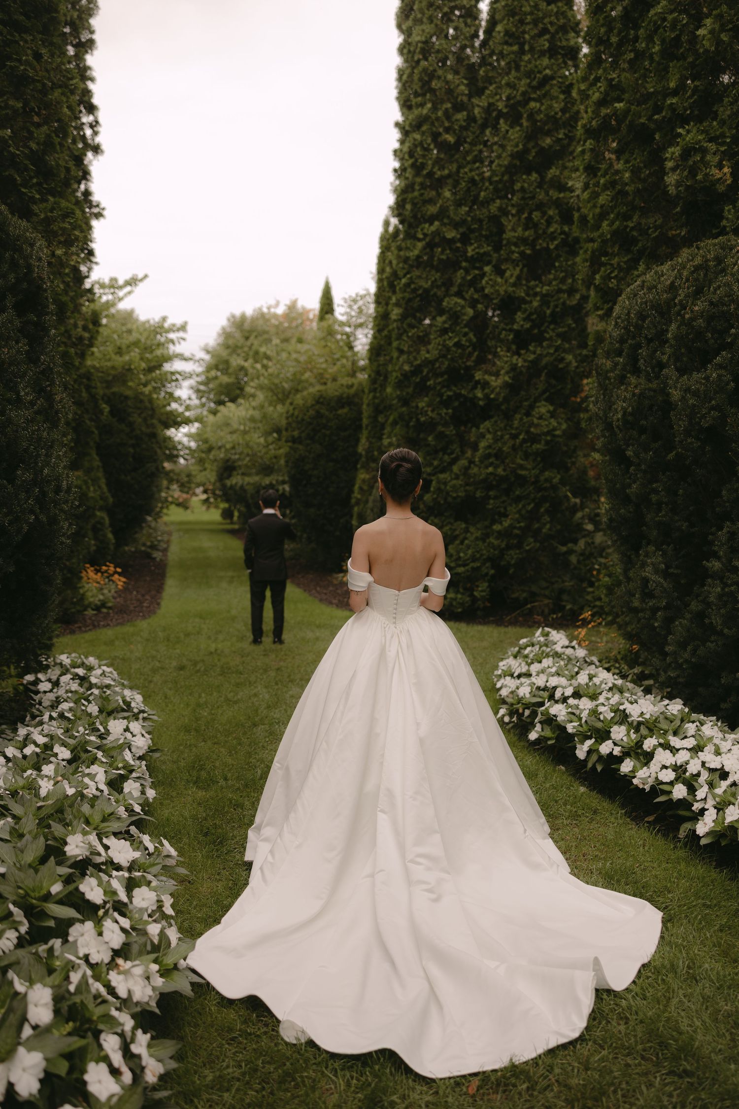 Bride in elegant off-shoulder white ball gown with long train walks toward groom in garden path.