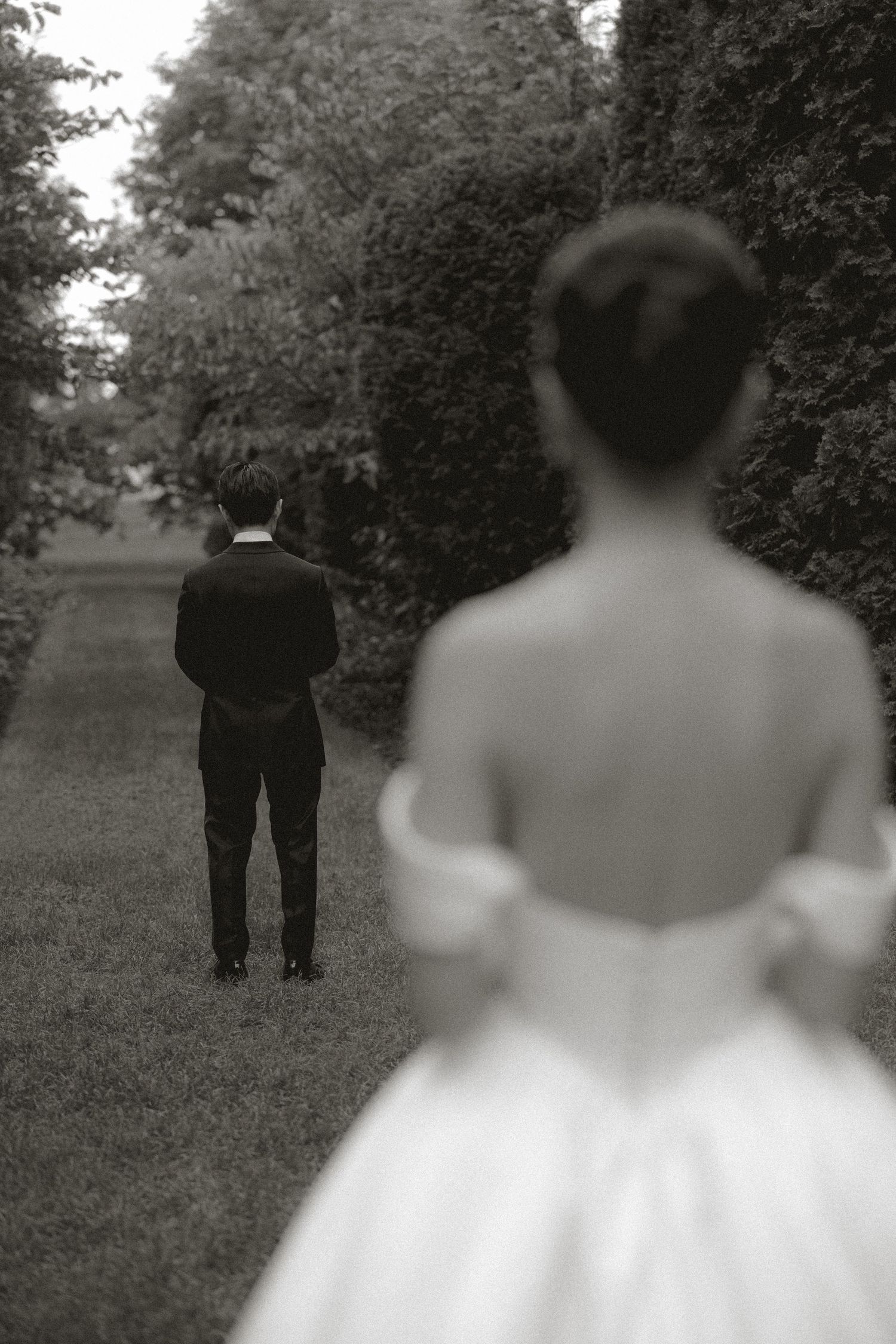 Black and white first look photo of groom standing ahead of bride in lush garden setting.