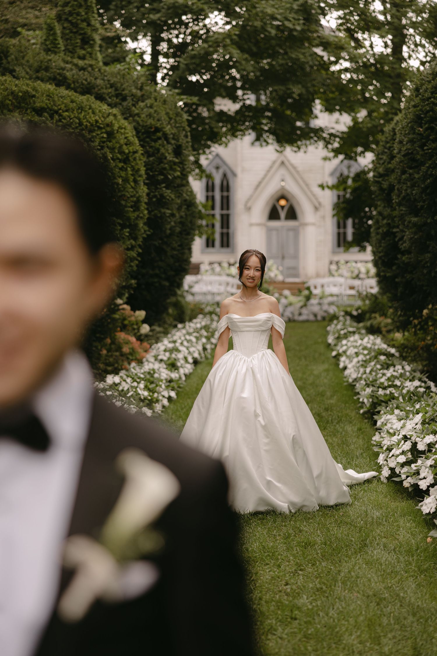 Bride in off-shoulder white ball gown walks toward groom along floral garden path near chapel.