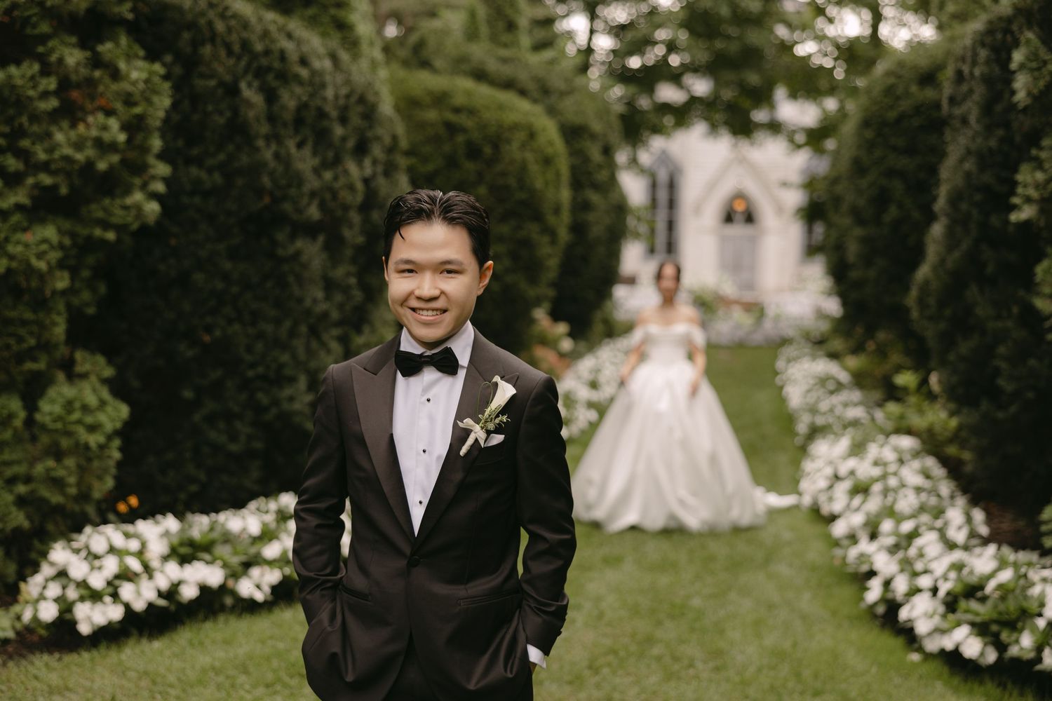 Smiling groom in black tuxedo poses in garden with bride in white ball gown in background.
