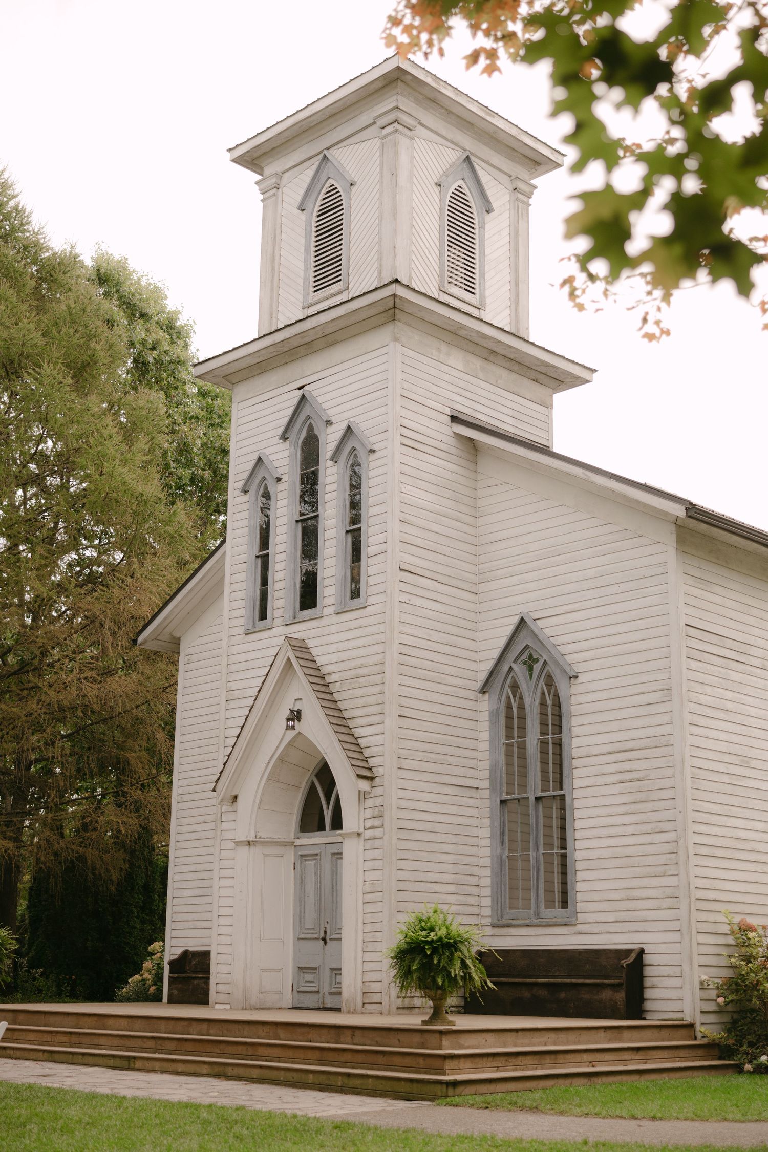 Charming white wooden country church with a bell tower and Gothic arched windows surrounded by trees.