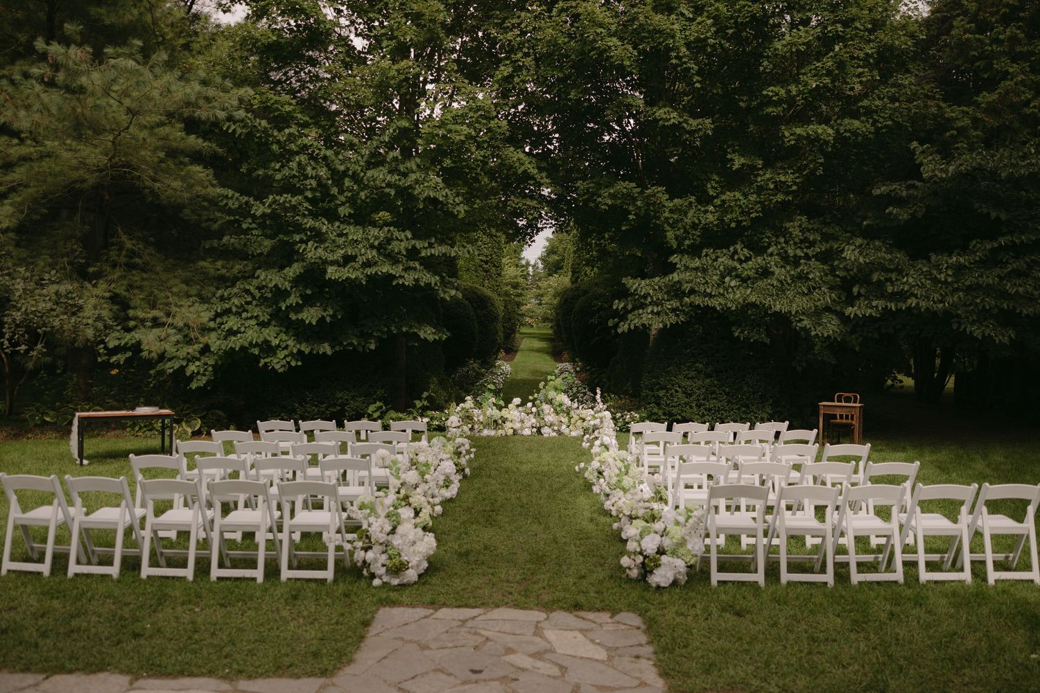 Outdoor wedding ceremony setup with white chairs lining a lush green garden aisle adorned with florals.