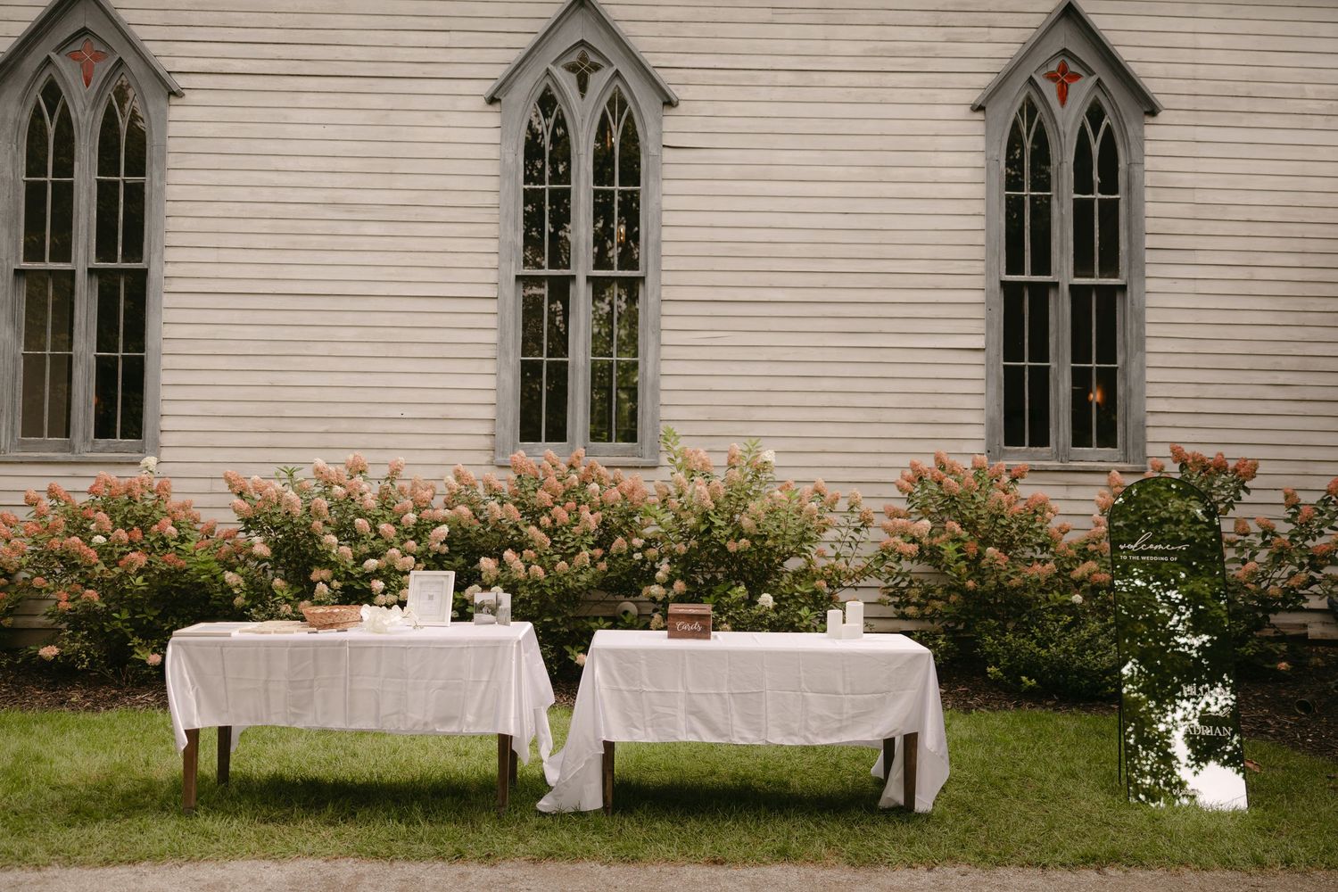 Two white linen-covered tables set outdoors beside a white wooden church surrounded by blooming shrubs.