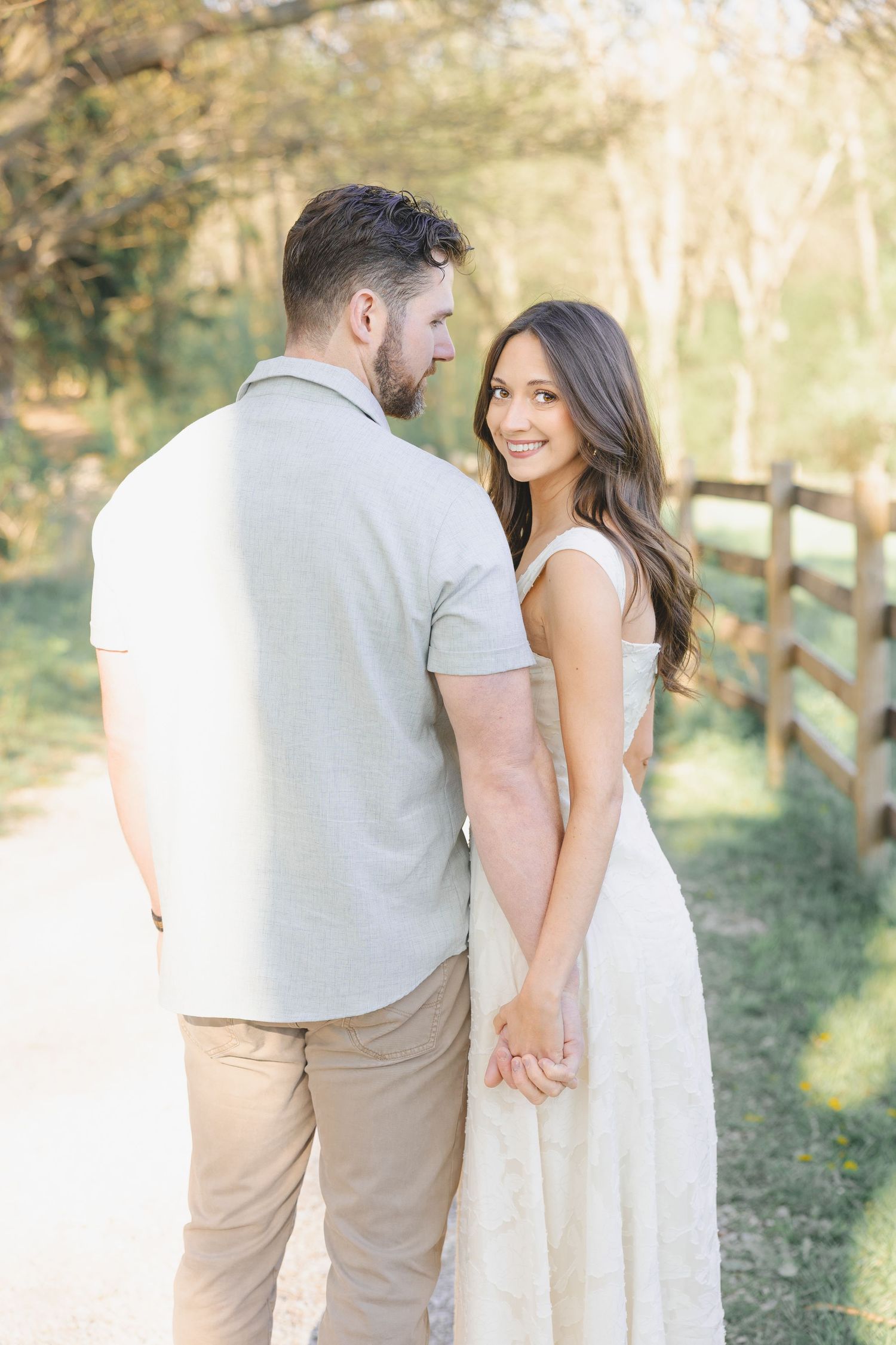 A couple holds hands on a sunlit path by a wooden fence, the woman in a white dress smiling over her shoulder.