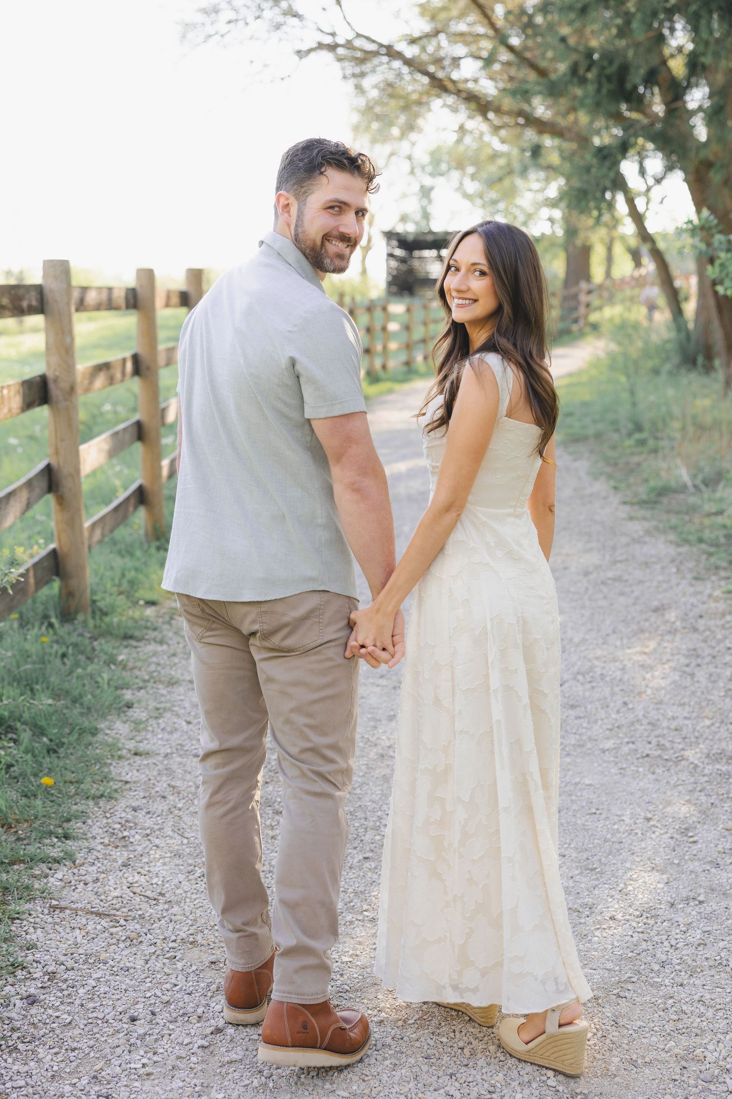 Couple holding hands on a rustic gravel path by a wooden fence, smiling back at camera in golden hour light.
