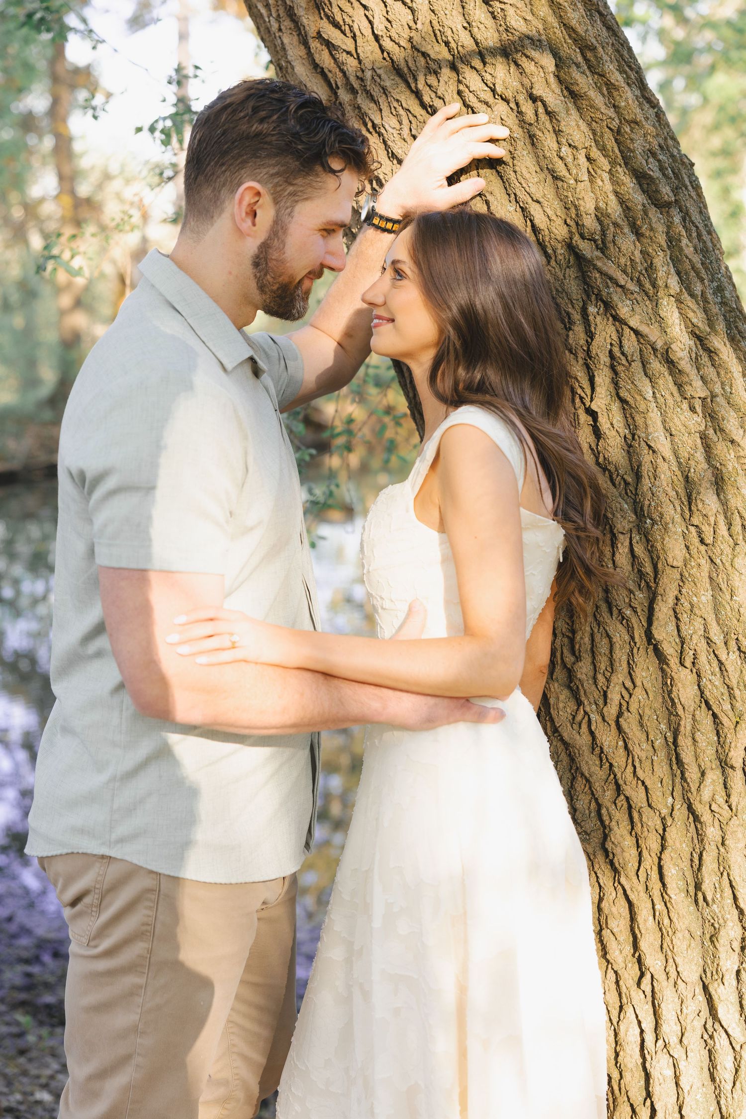 A romantic couple shares an intimate moment leaning against a tree in a sunlit forest, dressed in casual white and beige outfits.