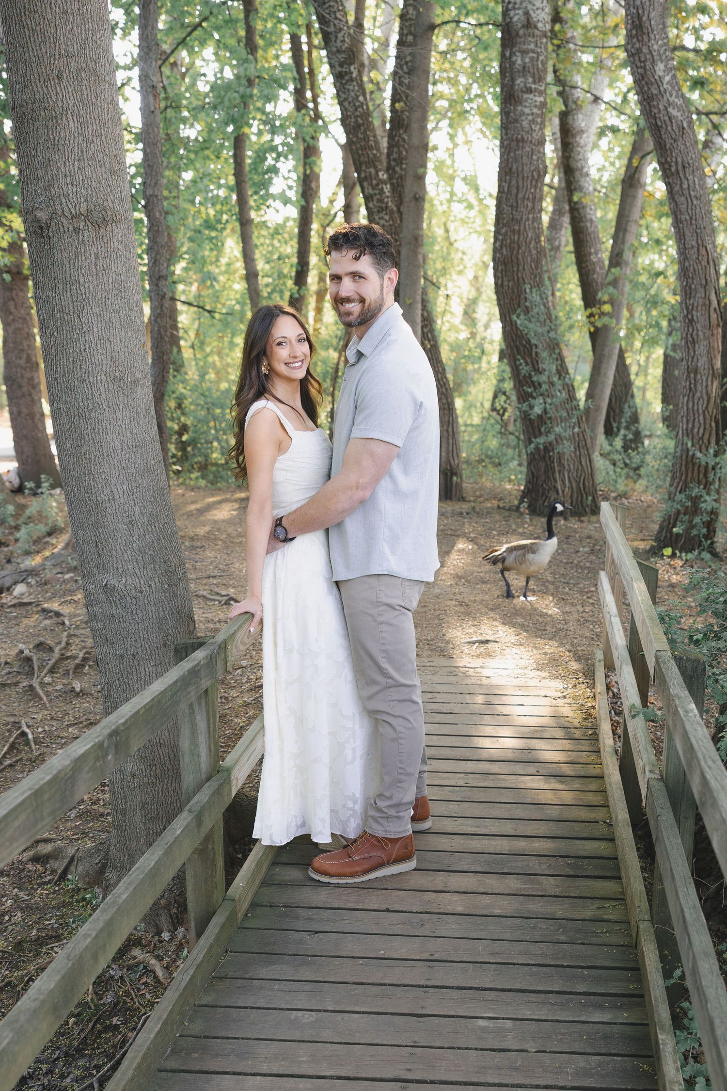 Couple embracing on a wooden boardwalk path surrounded by lush green trees in a forest setting.