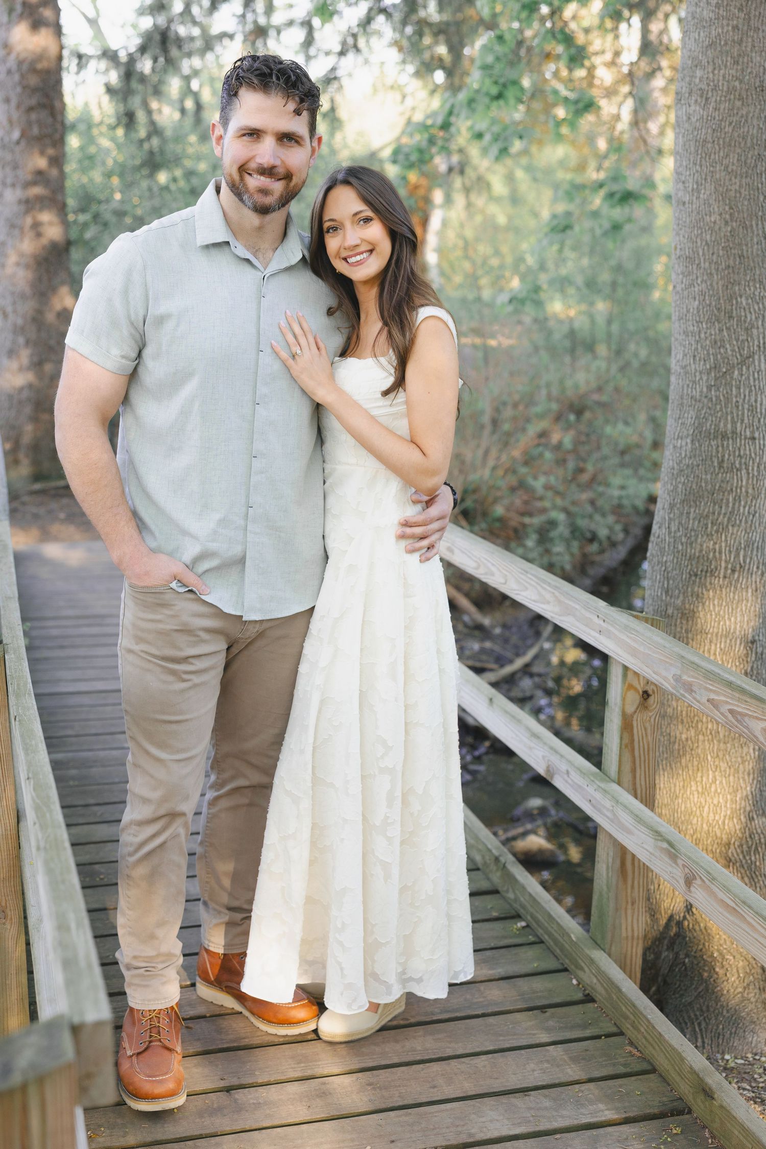 Smiling couple posing on a wooden bridge surrounded by trees, man in gray shirt and woman in white dress.
