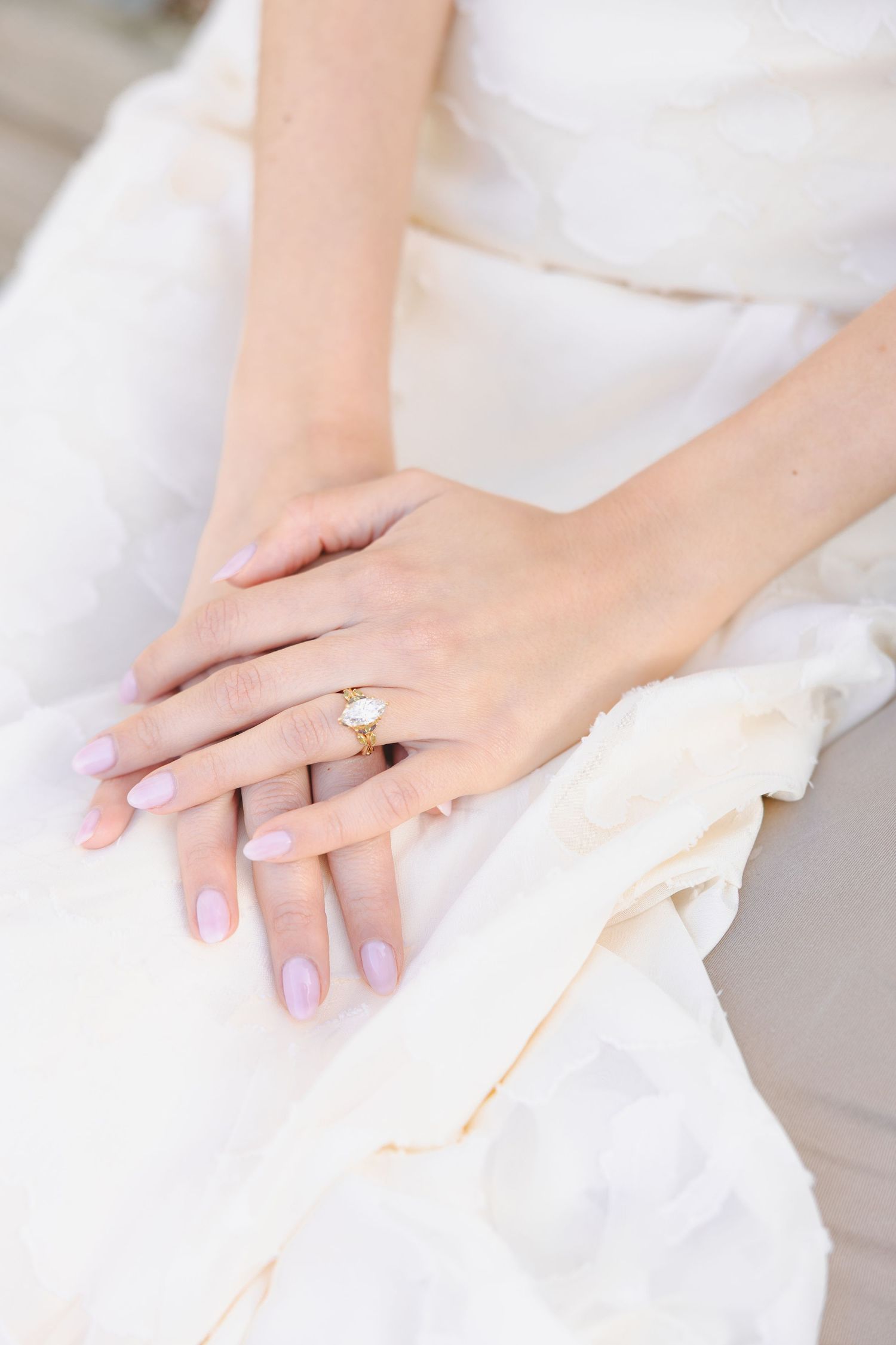Bride's hands resting on white wedding dress, wearing a gold solitaire engagement ring with light pink manicure.
