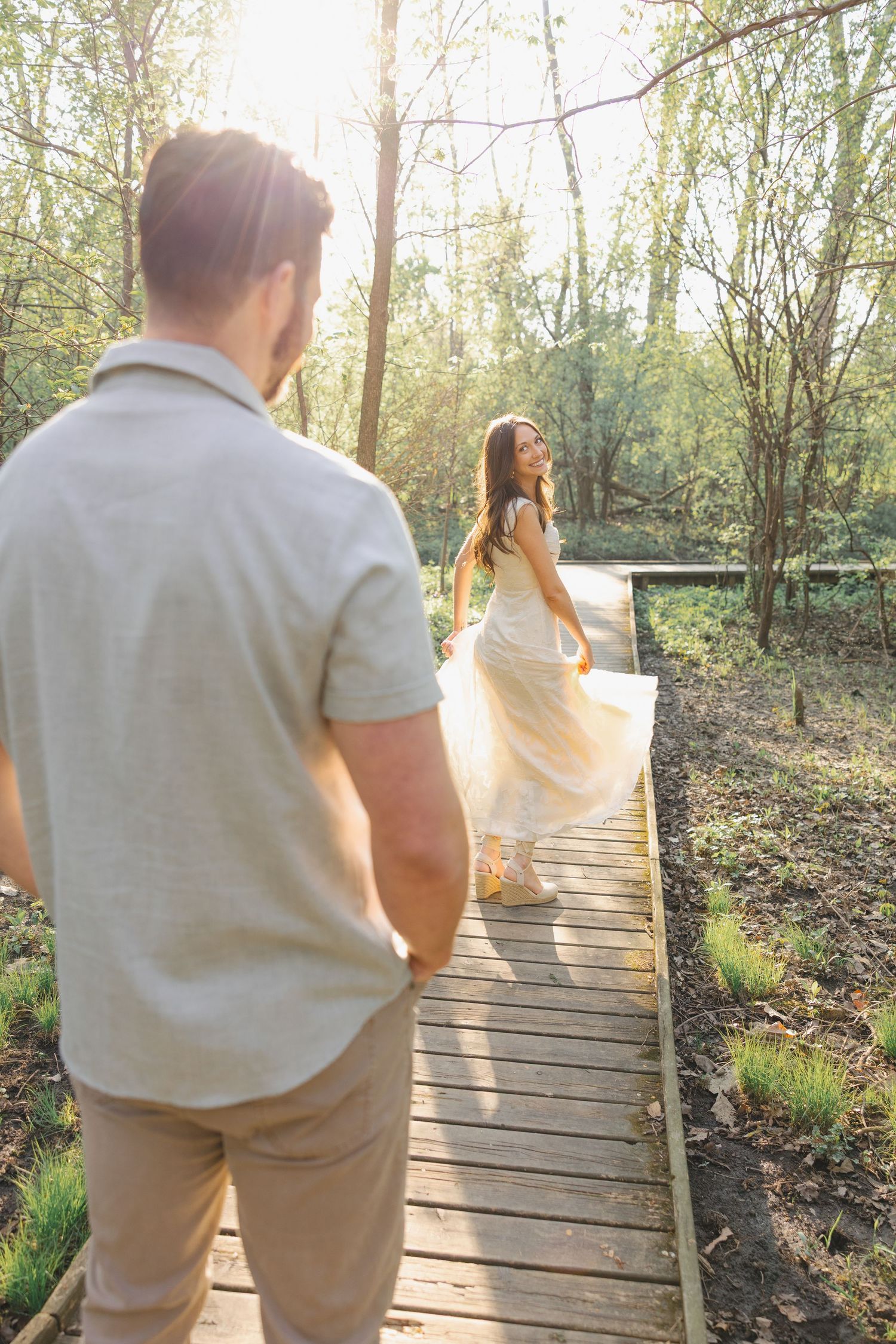 A couple walks on a wooden boardwalk in a sunlit forest, woman in flowing dress looks back smiling at man.