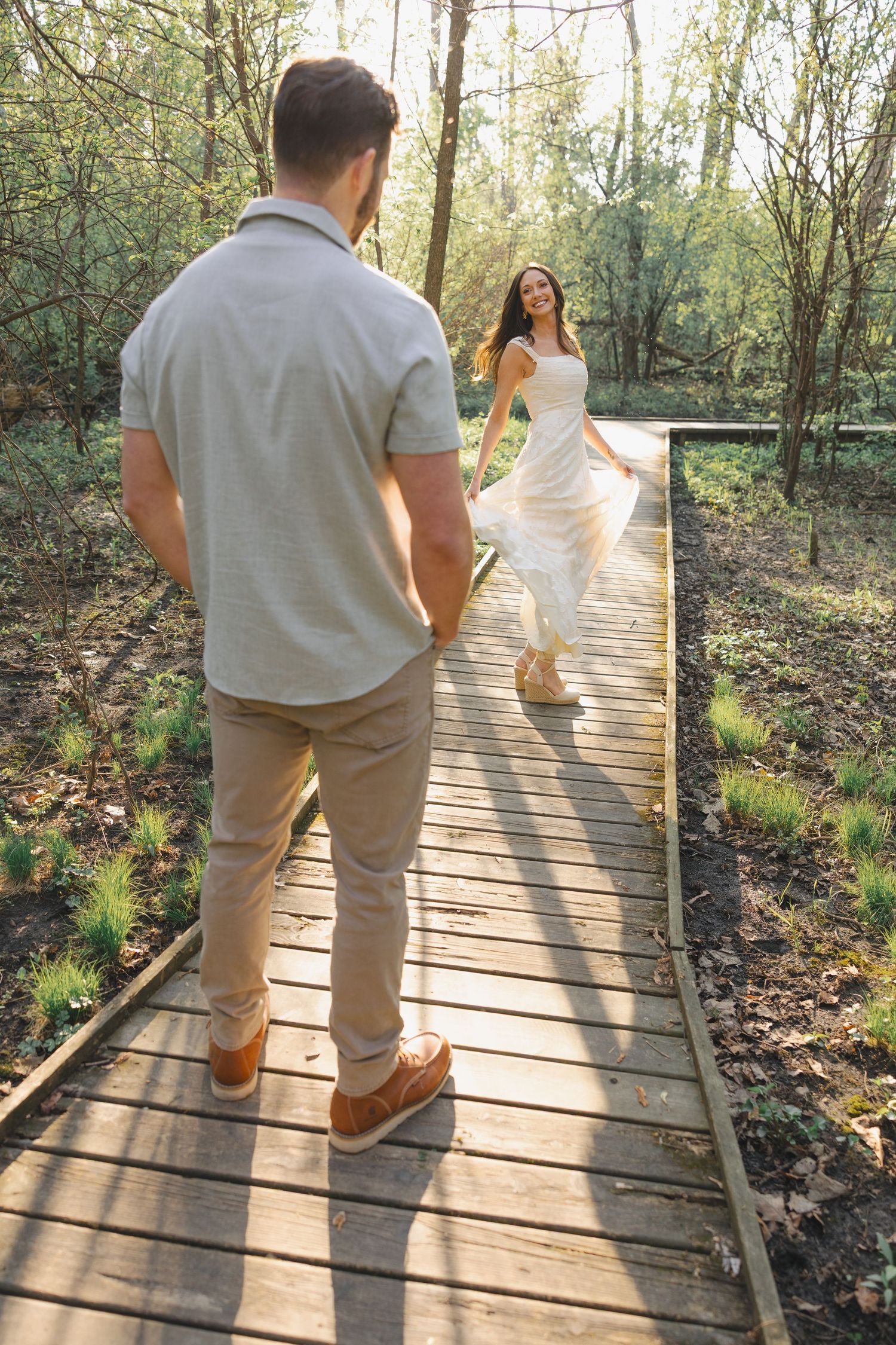 A couple on a wooden boardwalk in a forest, woman in white dress running toward man in casual attire.
