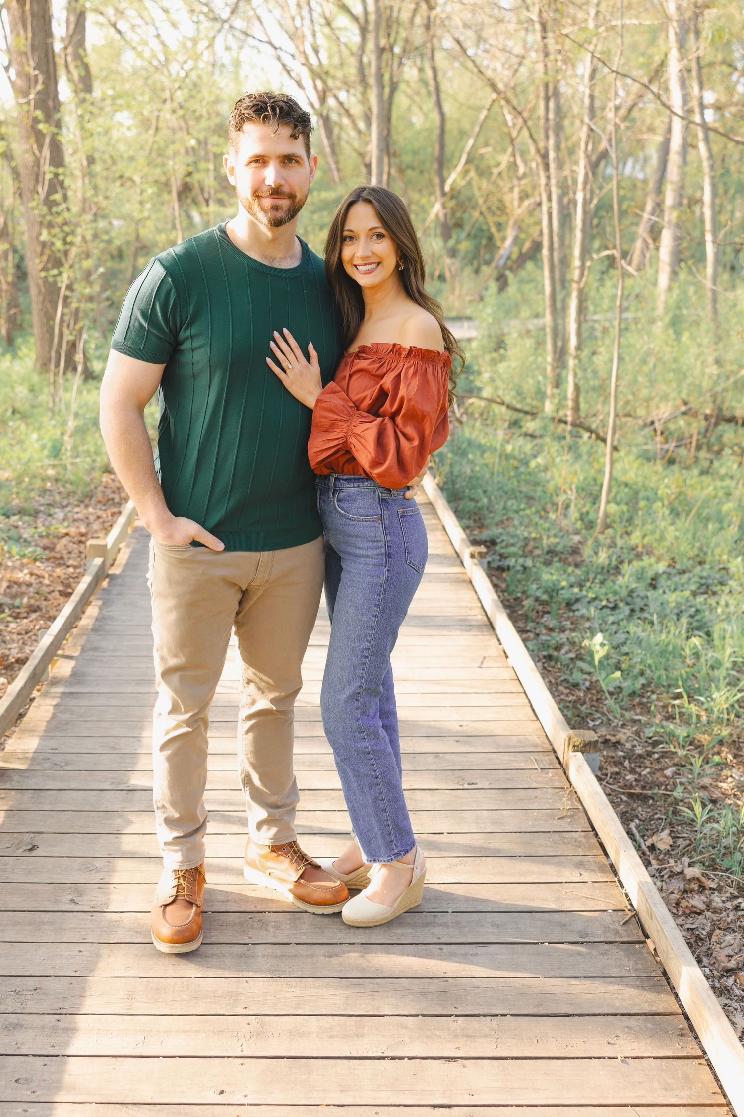 A smiling couple poses on a wooden boardwalk path surrounded by lush green trees in a park setting.
