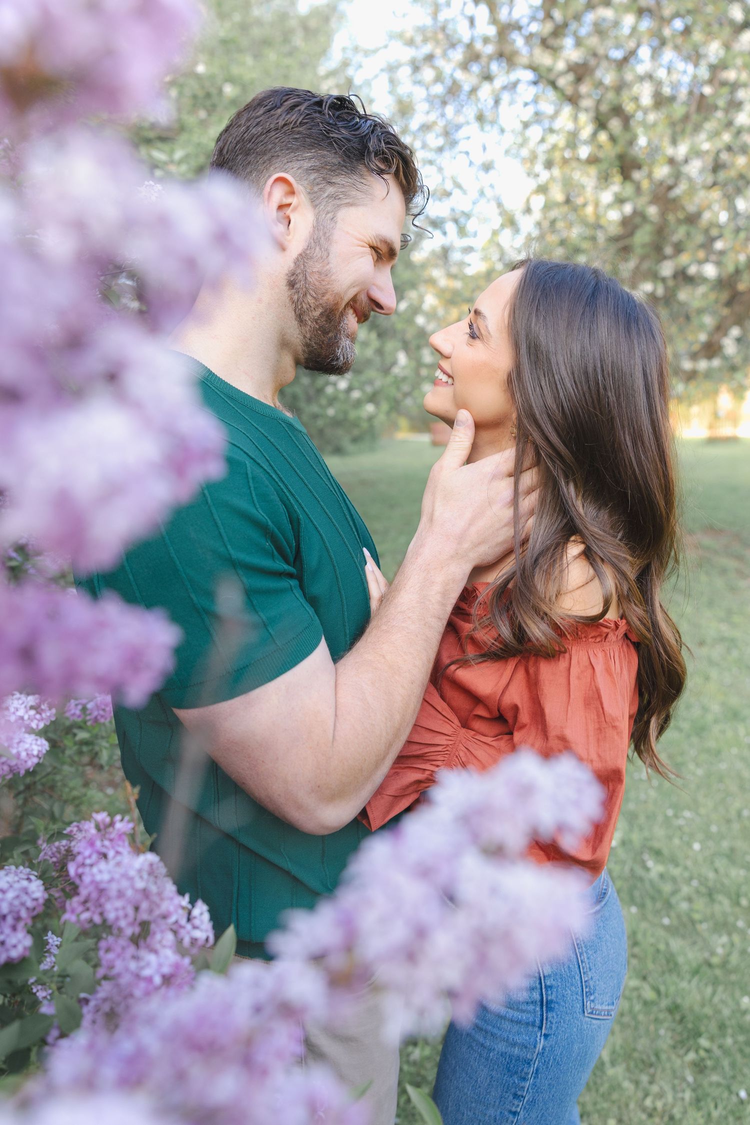 Couple gazing lovingly at each other surrounded by blooming lilac flowers in a lush green park setting.