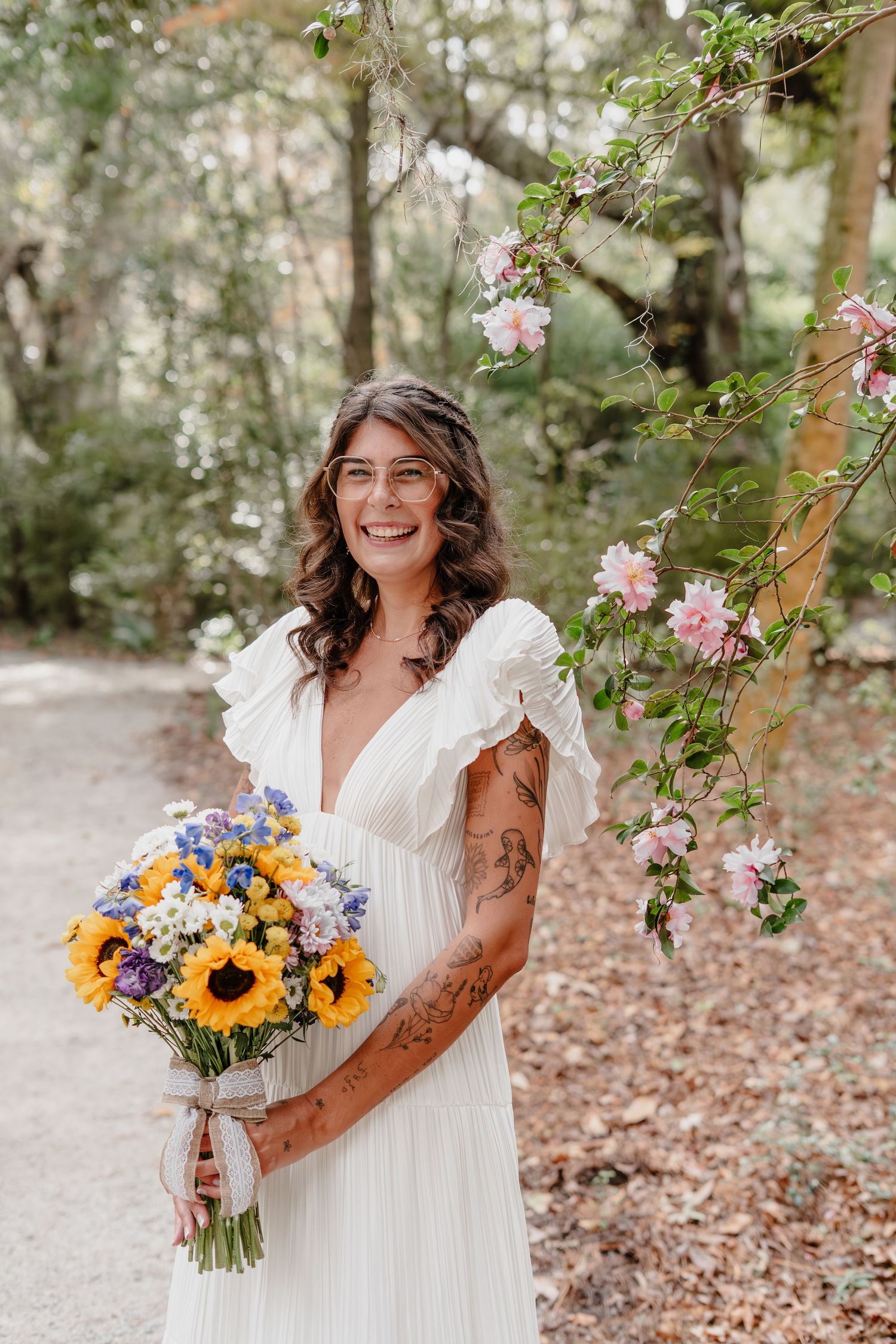 Smiling bride in white dress holding sunflower bouquet, standing near pink blossoms in a forest setting.