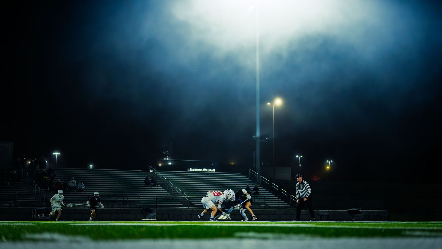 Two lacrosse players face off at midfield under stadium lights on a foggy night while a referee watches nearby at a Janesville game.