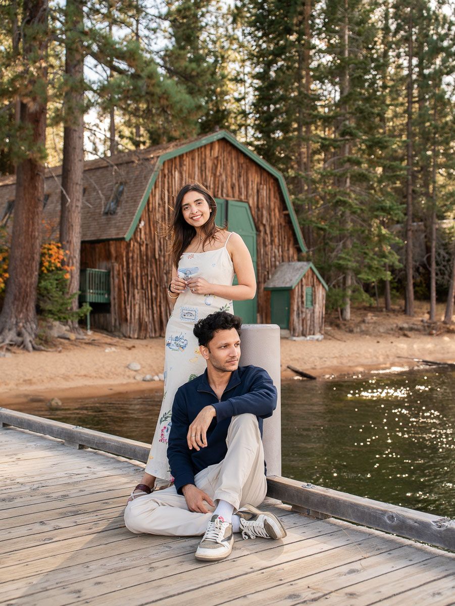 Couple posing on a wooden dock by a rustic log cabin near a lake surrounded by tall pine trees.