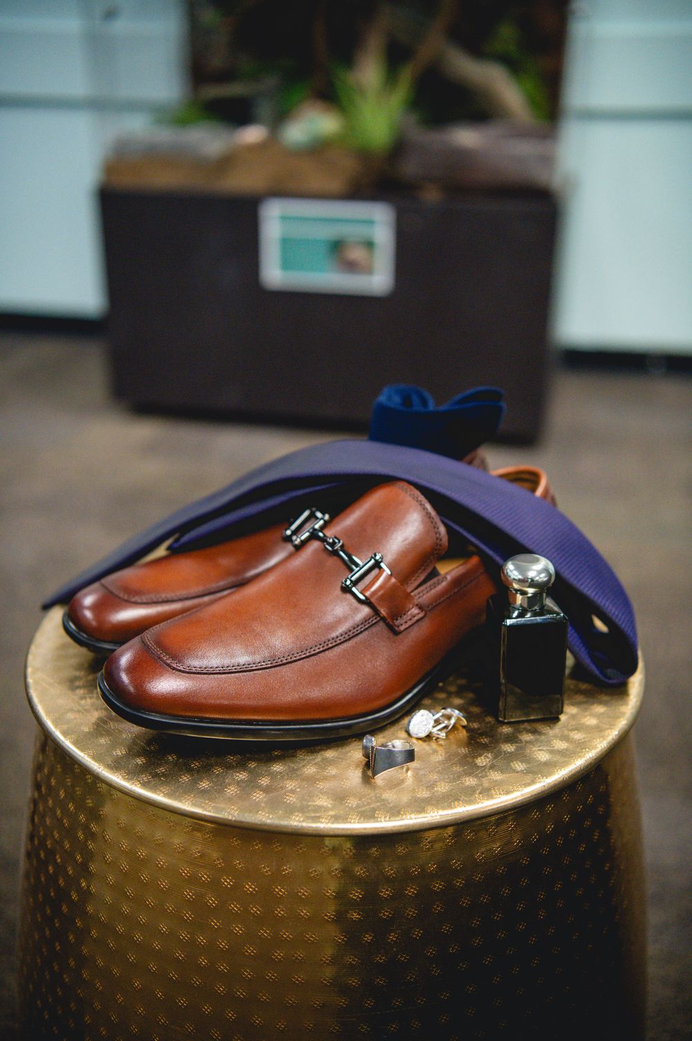 Brown leather loafers with silver buckle displayed on gold table with navy tie, cufflinks, and cologne bottle.