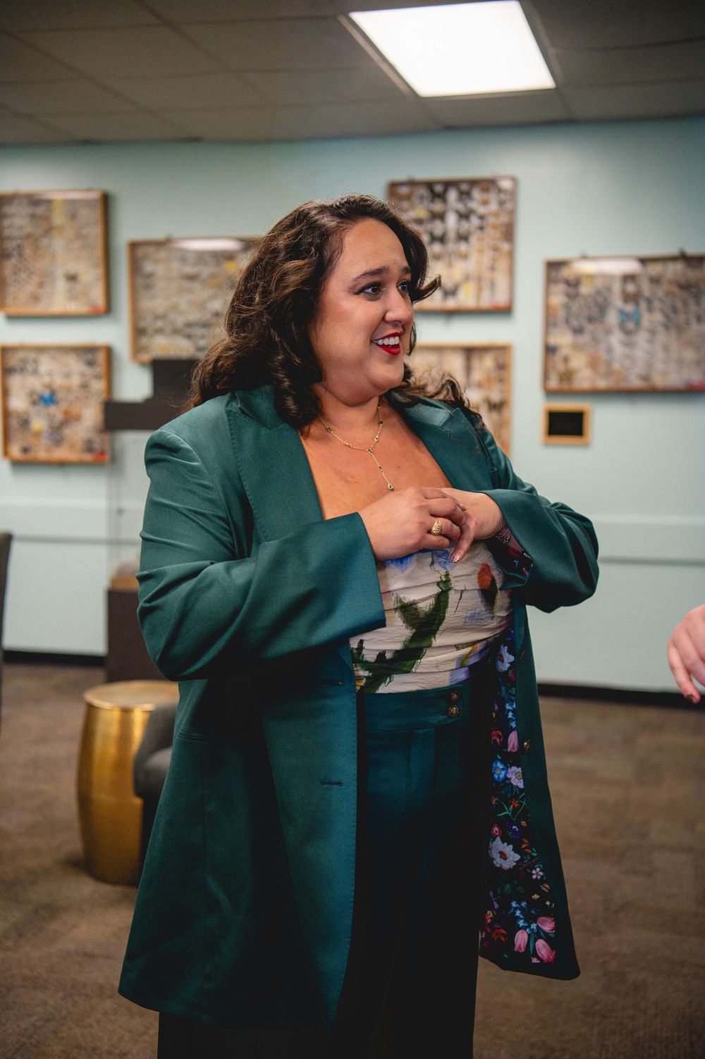 Woman in teal blazer and floral top smiling at an indoor gallery event with artwork on walls.