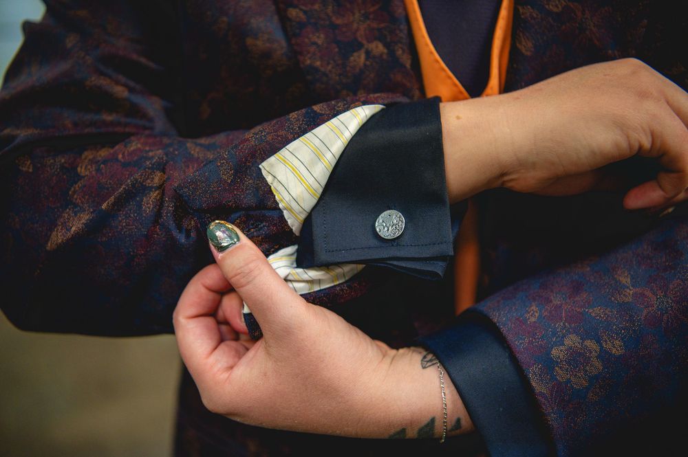 Person fastening a silver cufflink on a floral patterned suit jacket with an orange tie and tattooed hands.