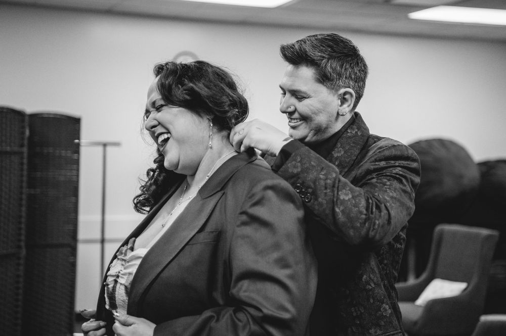 Black and white photo of a man helping a woman with her necklace, both smiling in an indoor setting.