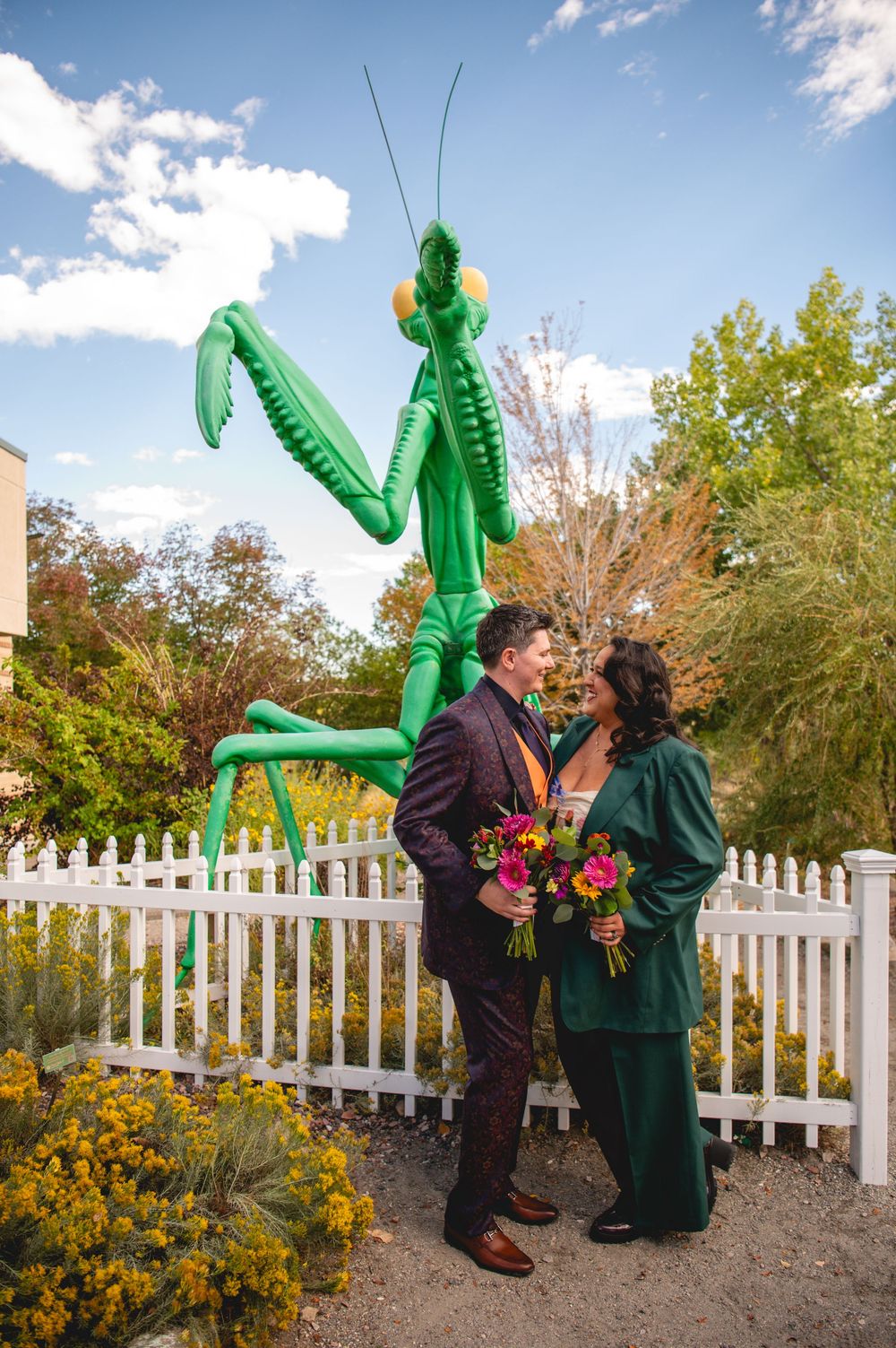 Couple poses romantically before giant green praying mantis sculpture by white picket fence, bride holds colorful bouquet.