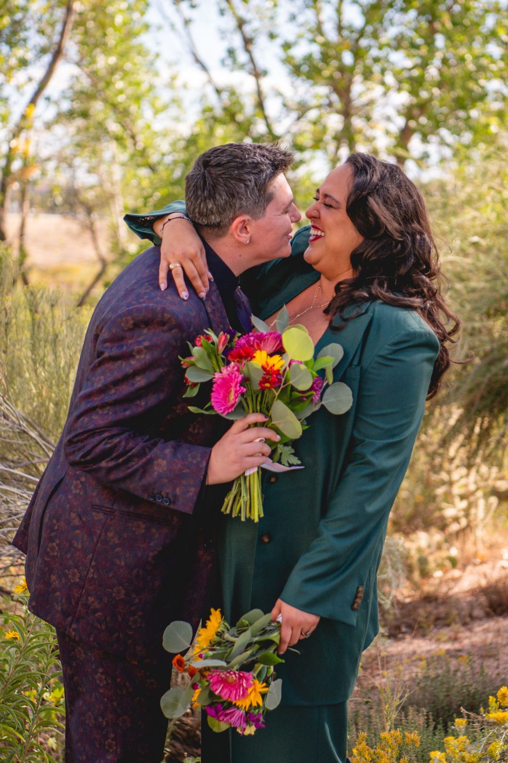 Couple in colorful formal attire sharing a joyful moment outdoors, holding vibrant pink and yellow bouquets among green trees.