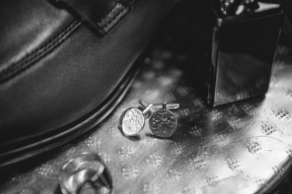 Black and white photo of elegant cufflinks beside a leather shoe and cologne bottle on a patterned surface.