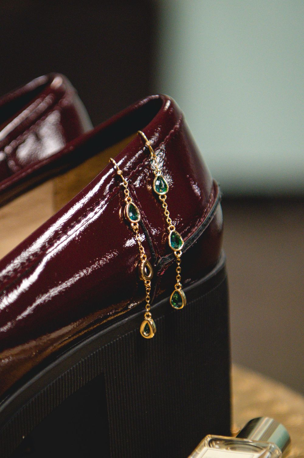 Gold chain earrings with emerald green gemstones resting against burgundy patent leather heels.