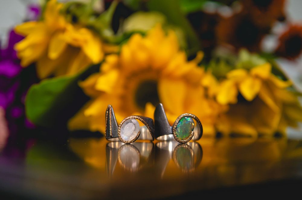Three ornate rings with gemstones displayed on a reflective surface with vibrant sunflowers in the background.