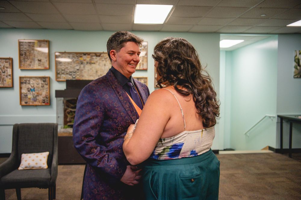 Couple in formal attire sharing a tender moment, man in purple suit, woman in teal dress, at an indoor event.