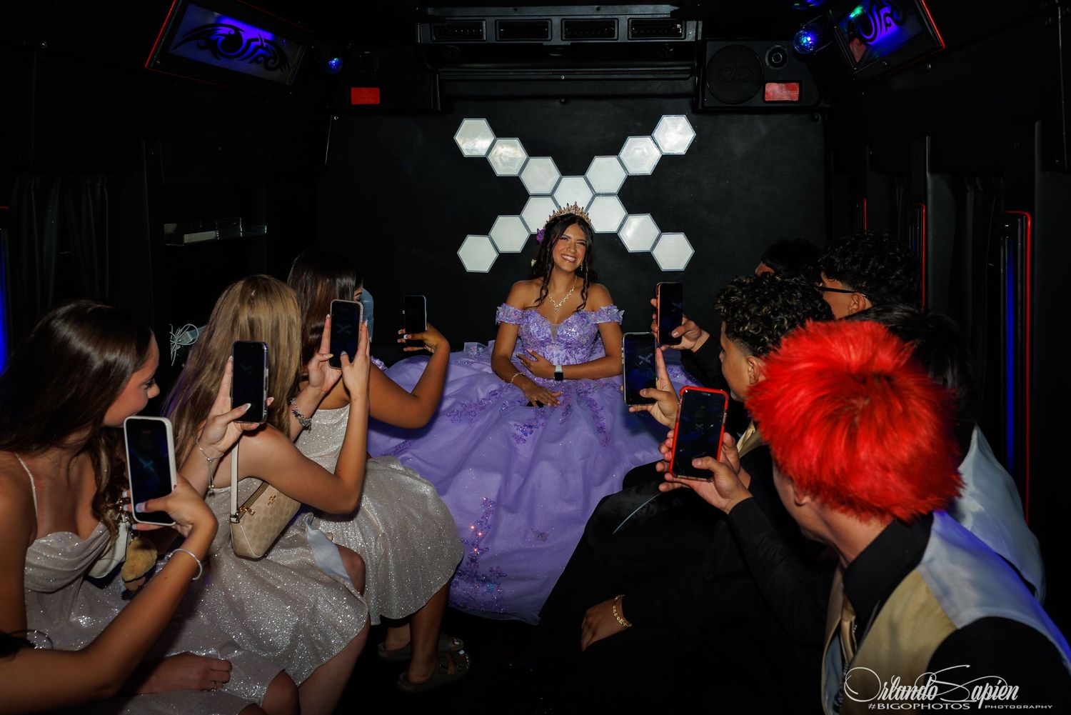 Teen girl in lavender quinceañera gown celebrated by friends with phones in a decorated party bus.