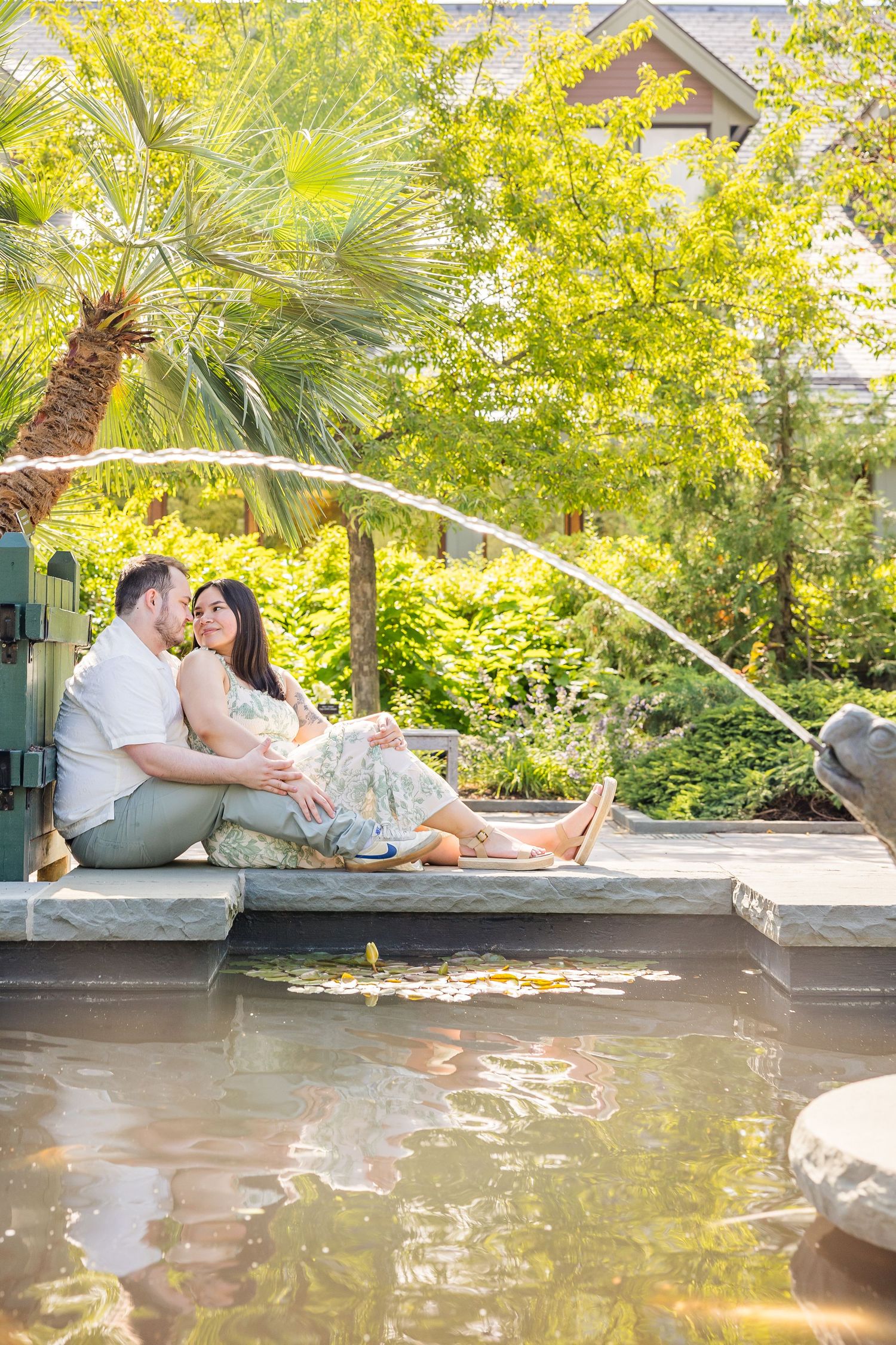 Couple sitting romantically by a garden fountain pond, surrounded by lush green trees and tropical palms on a sunny day.