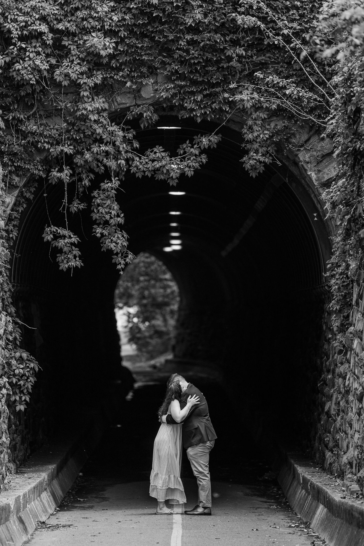 Black and white photo of a couple embracing and kissing inside an ivy-covered stone tunnel archway.