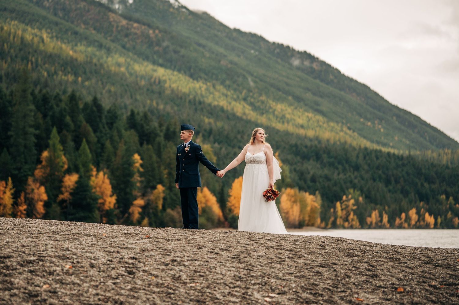 Bride and groom hold hands near a mountain lake surrounded by autumn foliage during an outdoor wedding ceremony.