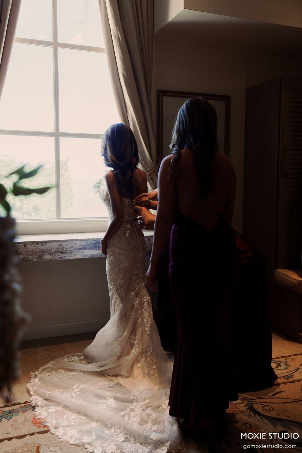 Bride in lace gown being helped by bridesmaid in purple dress near sunlit window on wedding day.