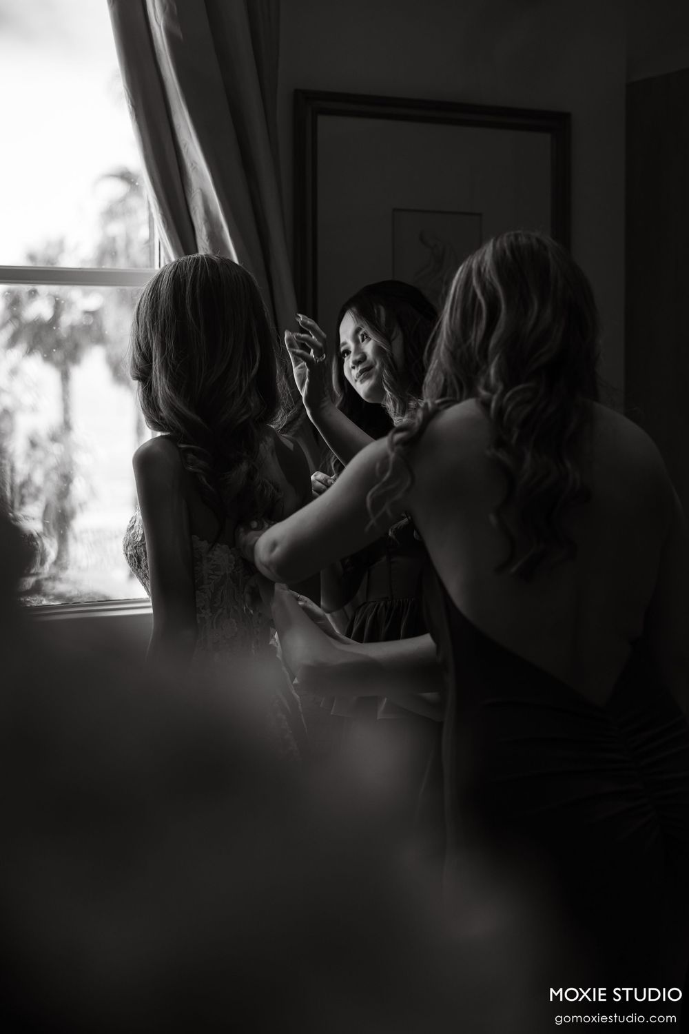 Black and white wedding photo of bride being helped by bridesmaids near a window, smiling in mirror reflection.