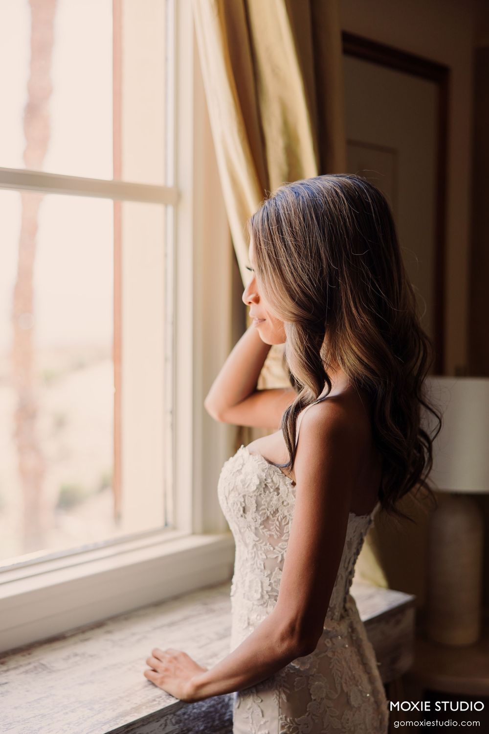 Bride in lace strapless gown gazing out a sunlit window, long brunette hair cascading over her shoulder.