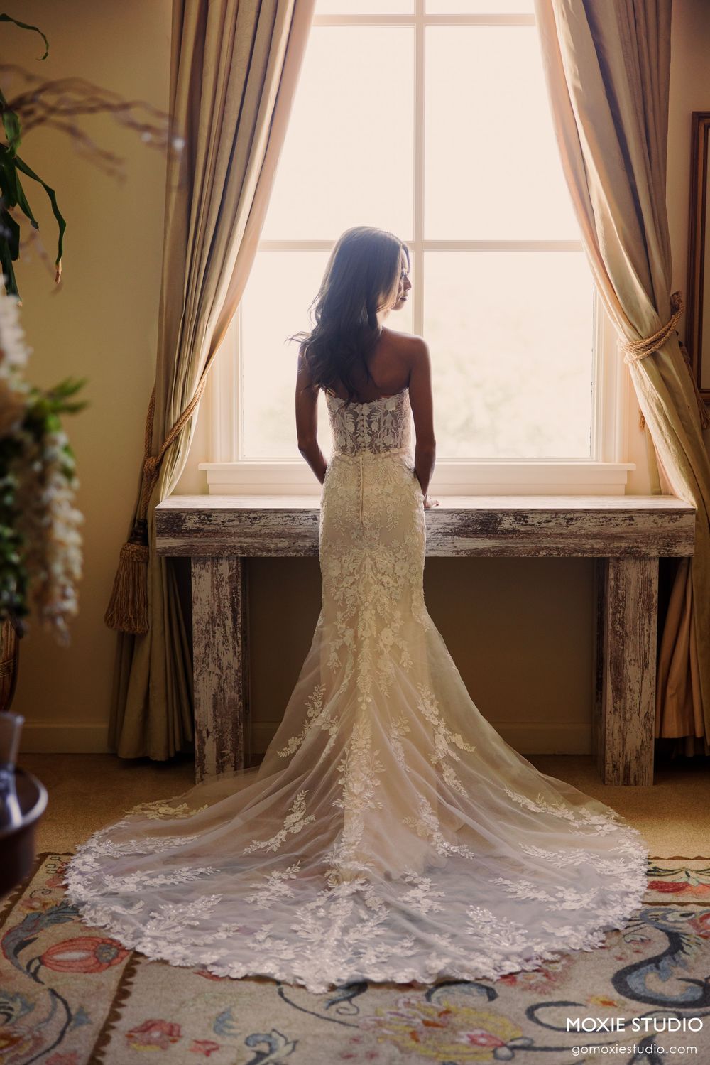 Bride in elegant lace mermaid wedding gown with train stands by sunlit window, photographed by Moxie Studio.