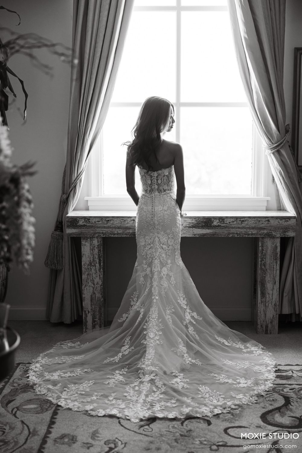 Bride in lace mermaid wedding gown with train stands at window in black and white portrait photo.
