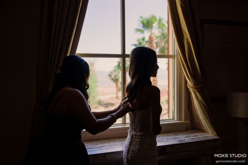 Silhouette of bride getting dressed by window with natural light, assisted by bridesmaid in elegant wedding preparation.