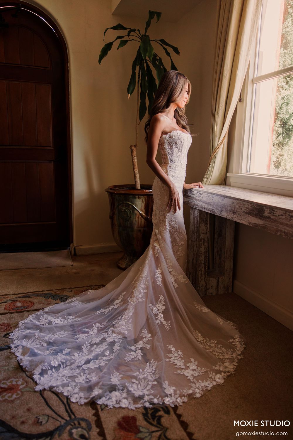 Bride in strapless lace mermaid wedding gown with long train stands by sunlit window in elegant room.