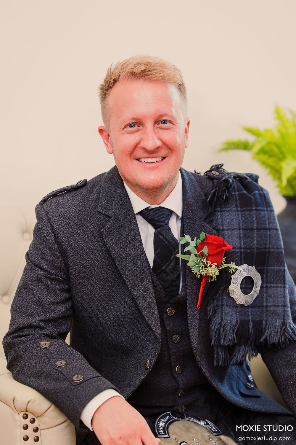 Smiling groom in Scottish tartan kilt outfit with red rose boutonniere, seated at wedding venue.