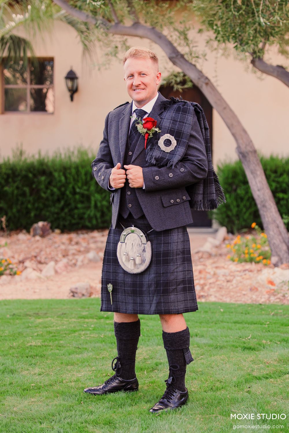 Man in traditional Scottish tartan kilt, jacket, and sporran poses outdoors on green lawn at a wedding.
