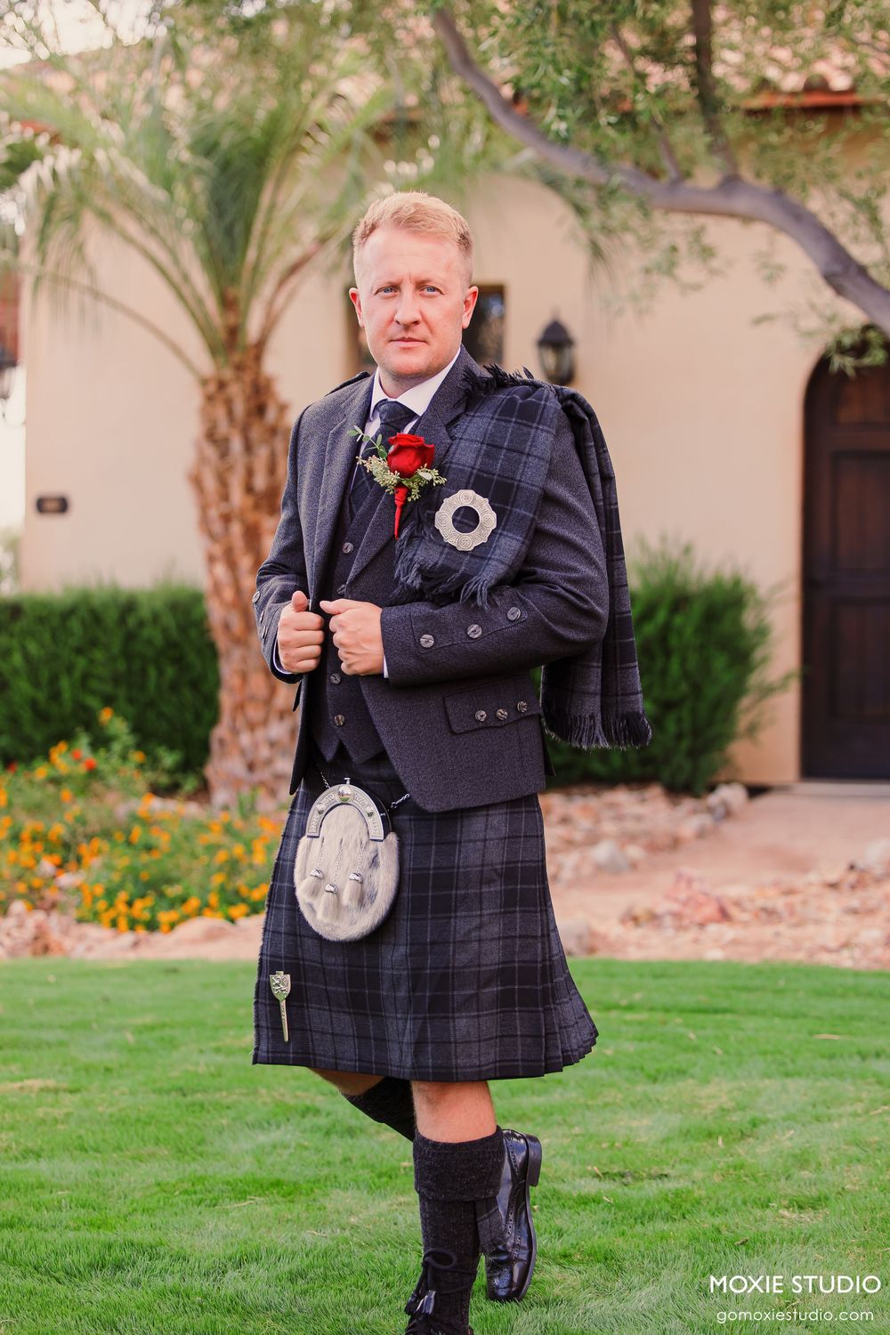 Man in traditional Scottish kilt attire with sporran and red boutonniere posing outdoors at a wedding.