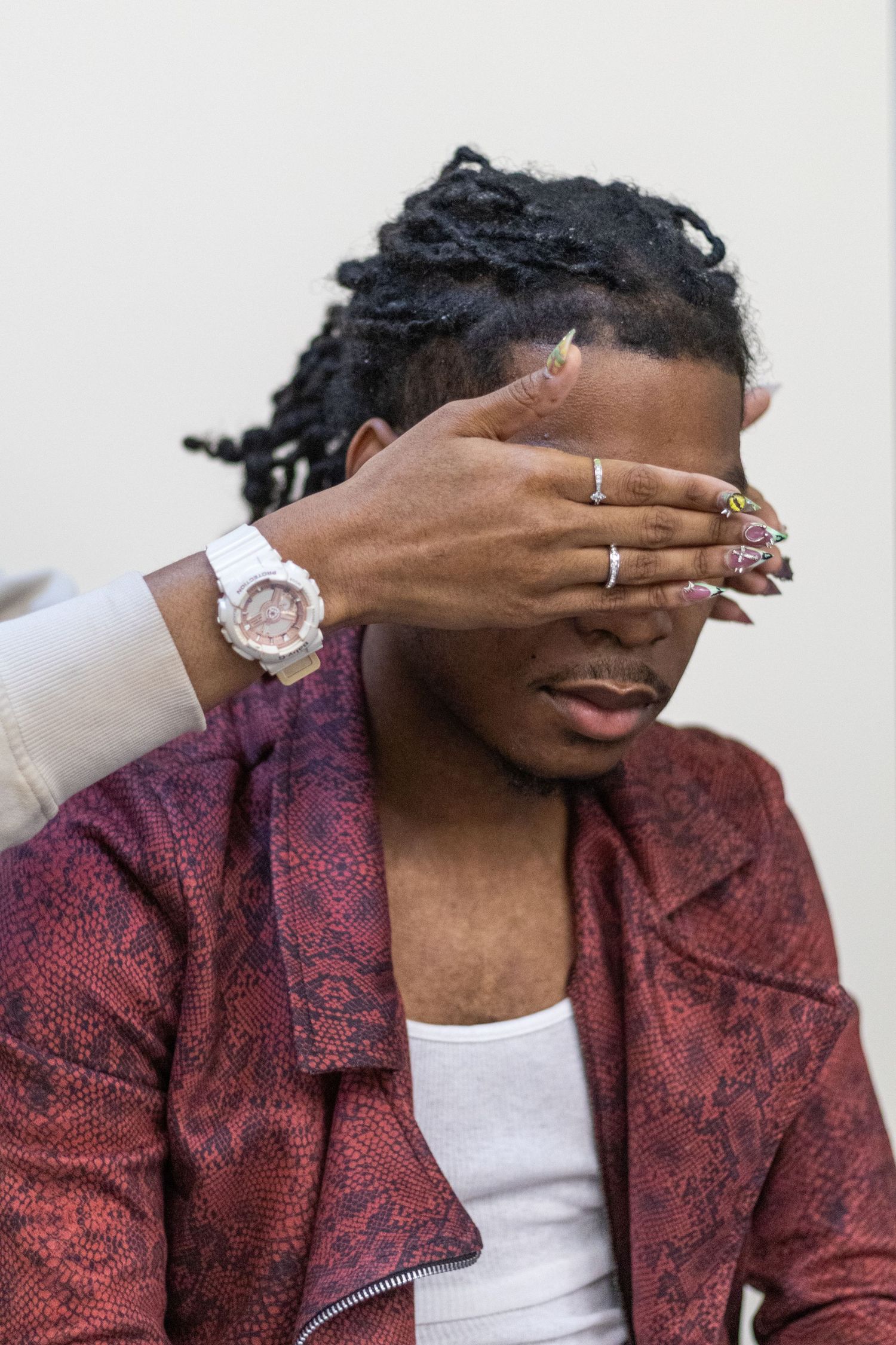A person covers another's face with a hand adorned with rings and long nails, wearing a white G-Shock watch.