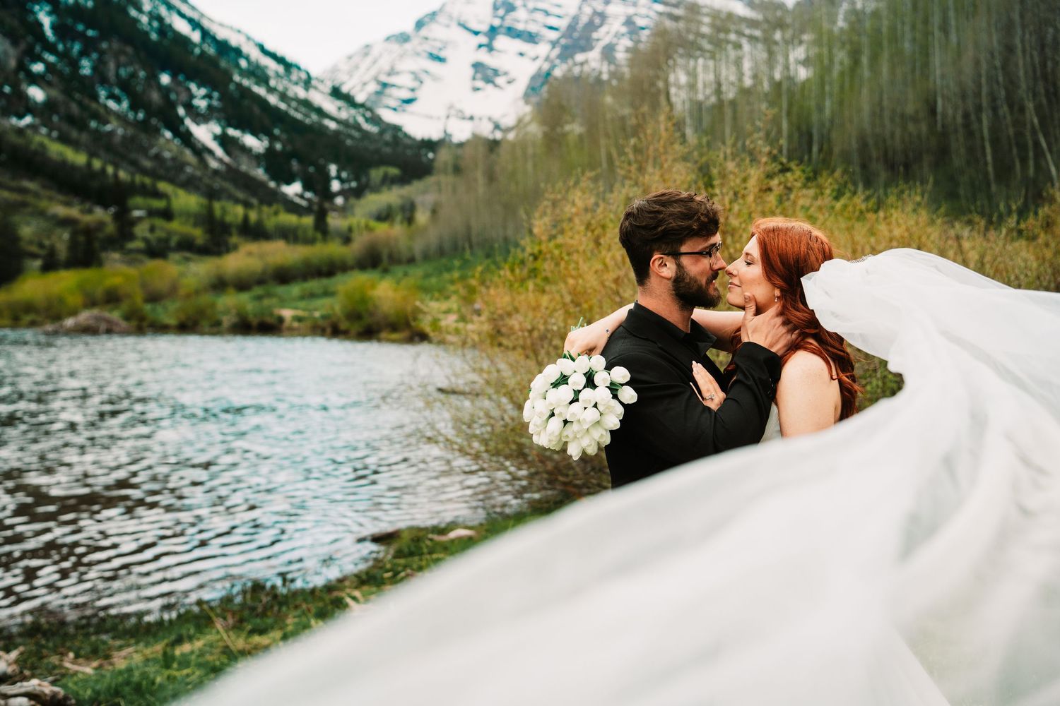 Bride and groom hold hands on rocky mountain stream surrounded by pine trees and snow-capped peaks during outdoor wedding.